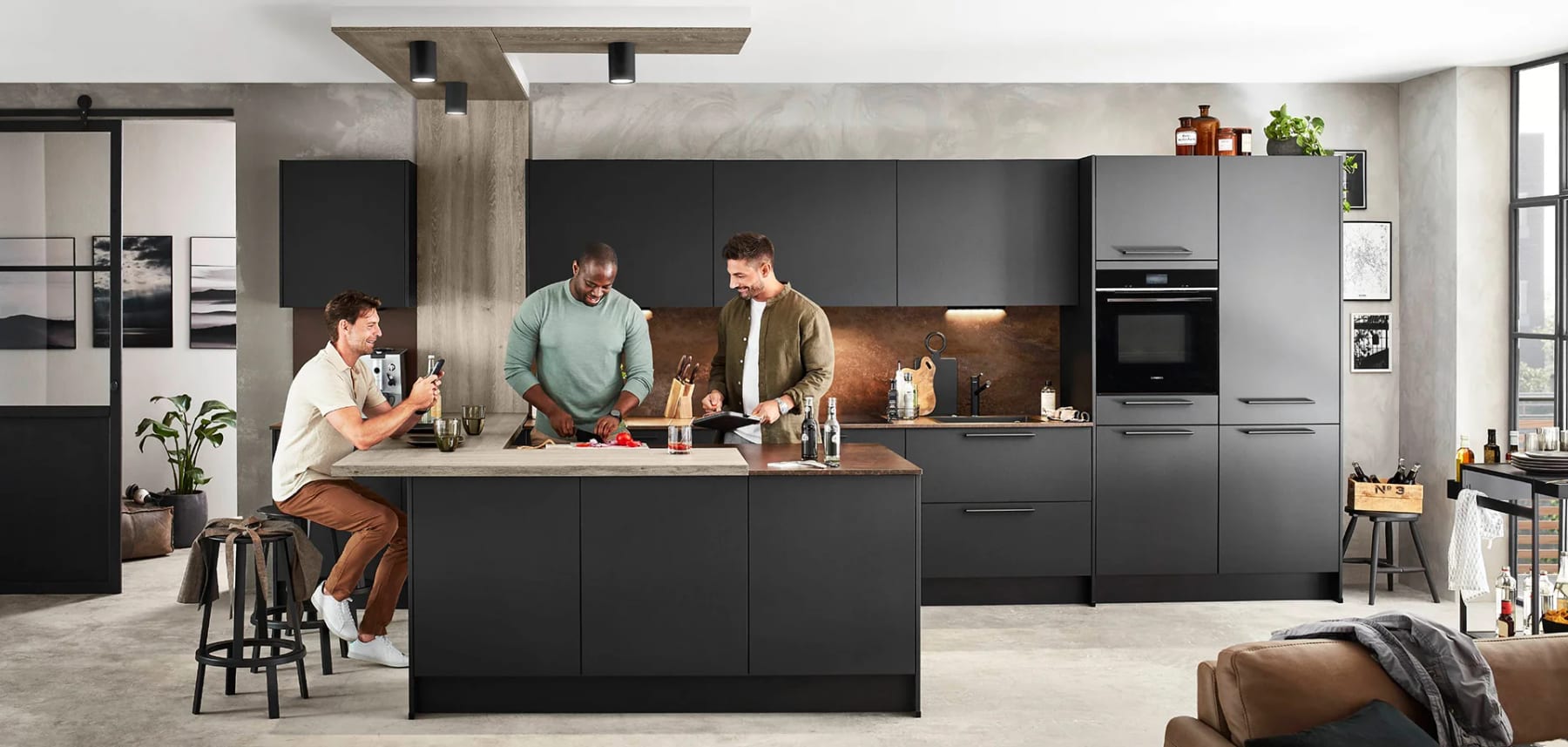 Cuisine ouverte aux façades noires mates, îlot et crédence cuivrée, trois personnes qui cuisinent