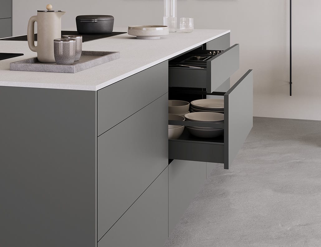 Bout d’un îlot gris anthracite, deux tiroirs ouverts l’un sous l’autre : ustensiles retenus par une tringle en haut, piles de bols et d’assiettes en bas