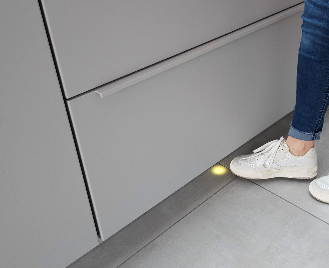 Un pied approche du socle d’un meuble bas ; le capteur logé dans la plinthe s’allume