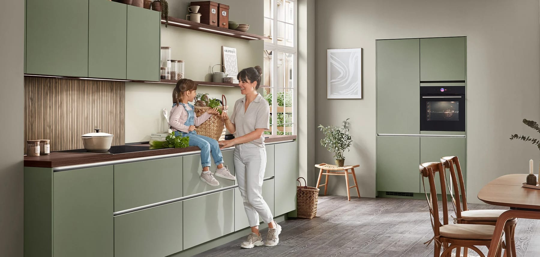 Cuisine linéaire aux façades vert olive mates, plan de travail et crédence en bois foncé, une mère et sa fille devant la table de cuisson