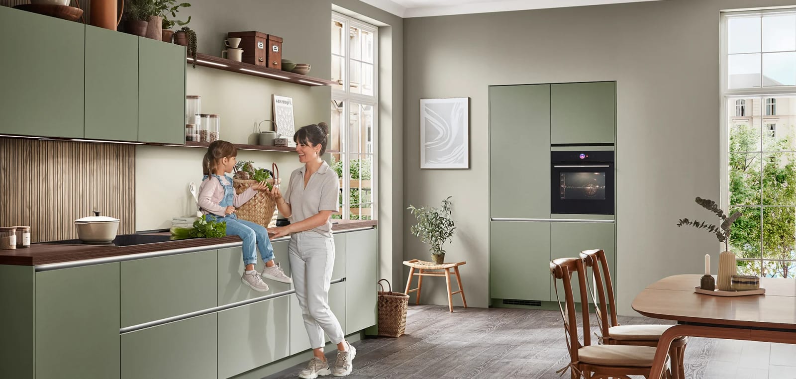 Cuisine en laque mate vert olive, plan de travail et crédence à lames de bois foncé, une mère et son enfant devant la fenêtre