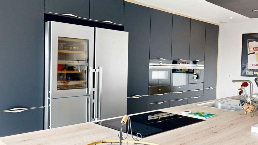 Cuisine posée aux façades anthracite mates : mur de colonnes toute hauteur, réfrigérateur à cave à vin, fours et machine à café encastrés à hauteur d’homme, îlot au plan décor chêne clair avec plaque à induction
