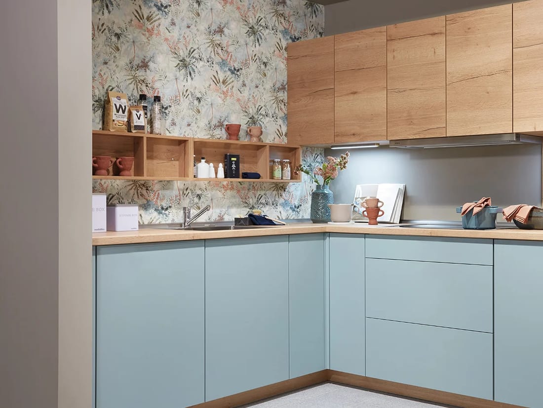 Cuisine d’angle en L à façades bleu aqua super-mates, meubles hauts en décor chêne, papier peint fleuri au-dessus du plan