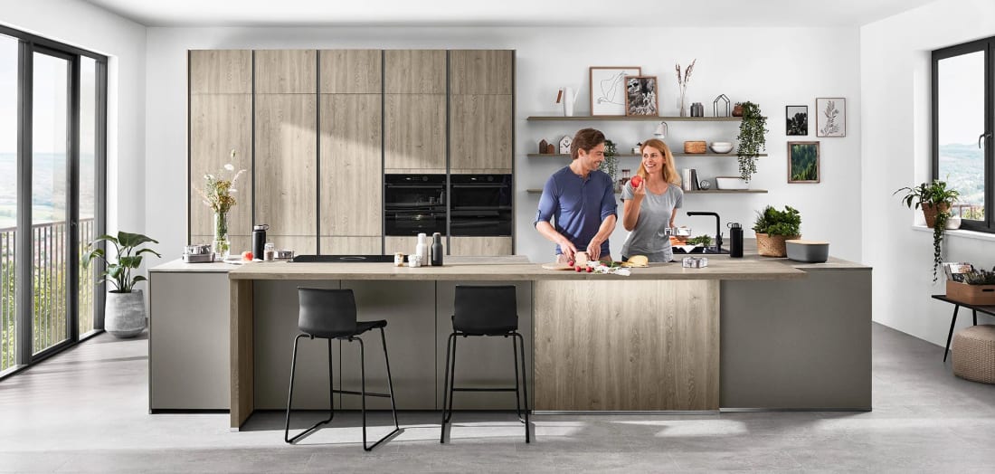 Cuisine à îlot en décor chêne au veinage vertical, façades hautes sans poignée et colonne de fours intégrée à l’alignement