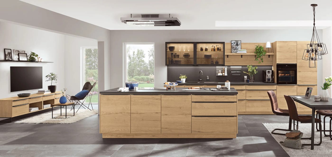 Cuisine ouverte en décor chêne clair : îlot au plan de travail sombre, coin télévision d’un côté, table à manger de l’autre