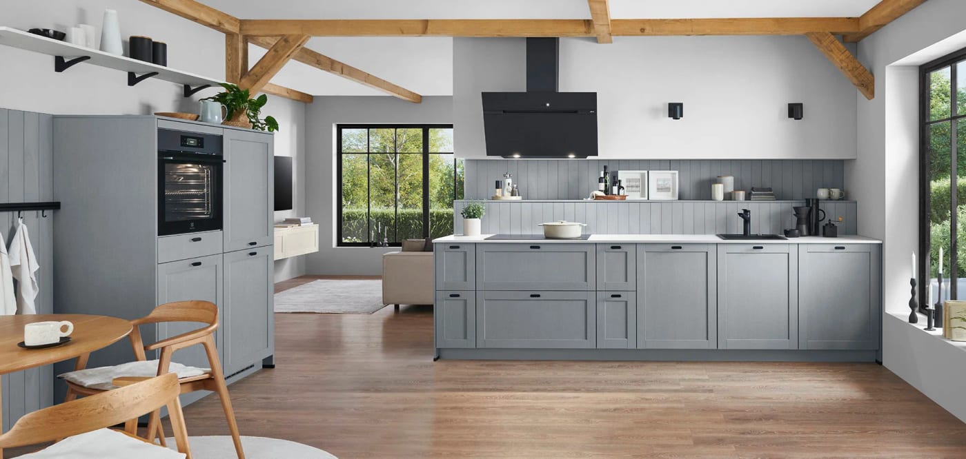 Cuisine à cadre gris bleuté sous des poutres apparentes, ouverte sur le séjour d’une maison de campagne